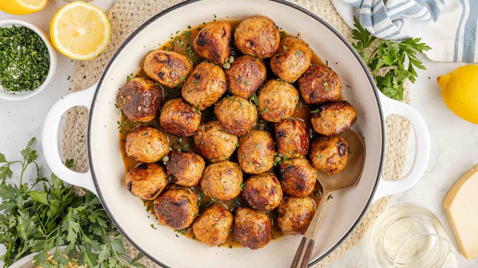 A white dish filled with browned meatballs in sauce, garnished with chopped herbs, with a serving spoon and fresh ingredients like lemon and parsley around it.
