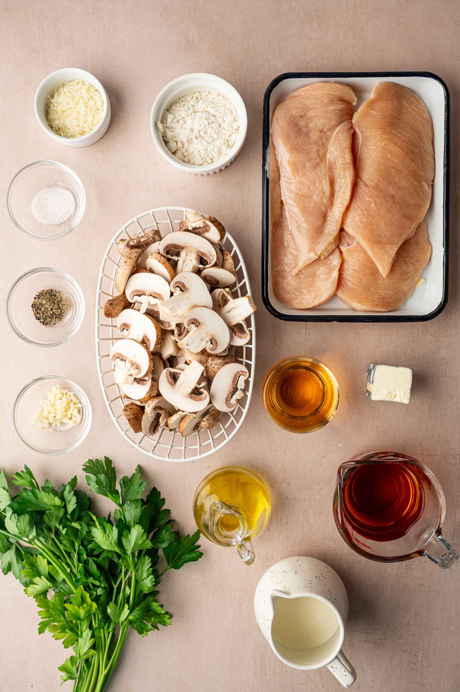 Overhead view of raw chicken breasts, sliced mushrooms, fresh parsley, and various chicken marsala ingredients in bowls including oil, butter, cream, broth, garlic, cheese, flour, salt, and pepper.