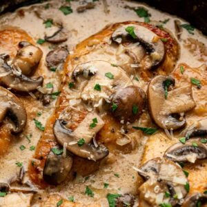 Chicken breasts cooked in a creamy sauce with sliced mushrooms and garnished with chopped parsley in a skillet.