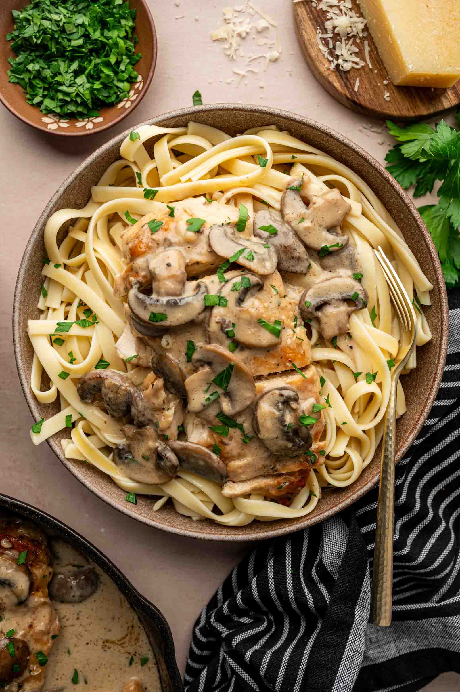A bowl of fettuccine pasta topped with creamy chicken marsala, garnished with chopped parsley, with a fork on the side. Parmesan cheese and fresh herbs are nearby.