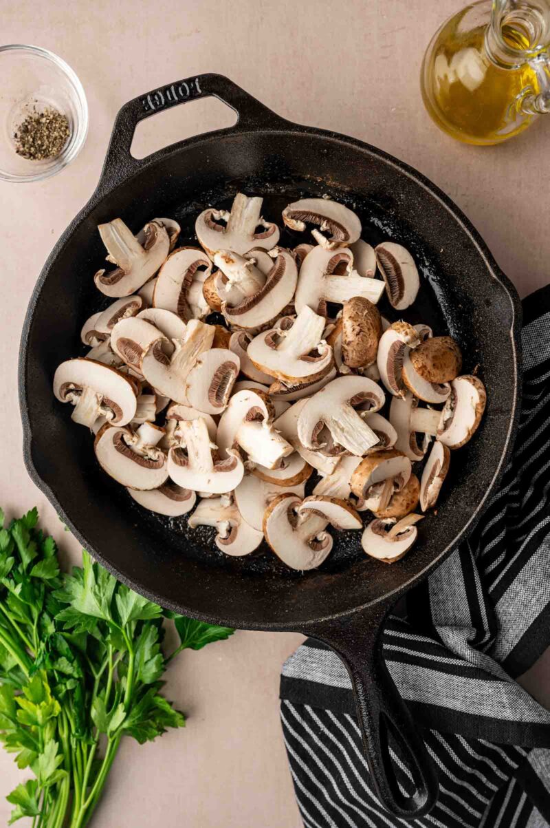Sliced mushrooms in a black cast iron skillet, with a small bowl of pepper, a bottle of oil, parsley, and a striped kitchen towel nearby.