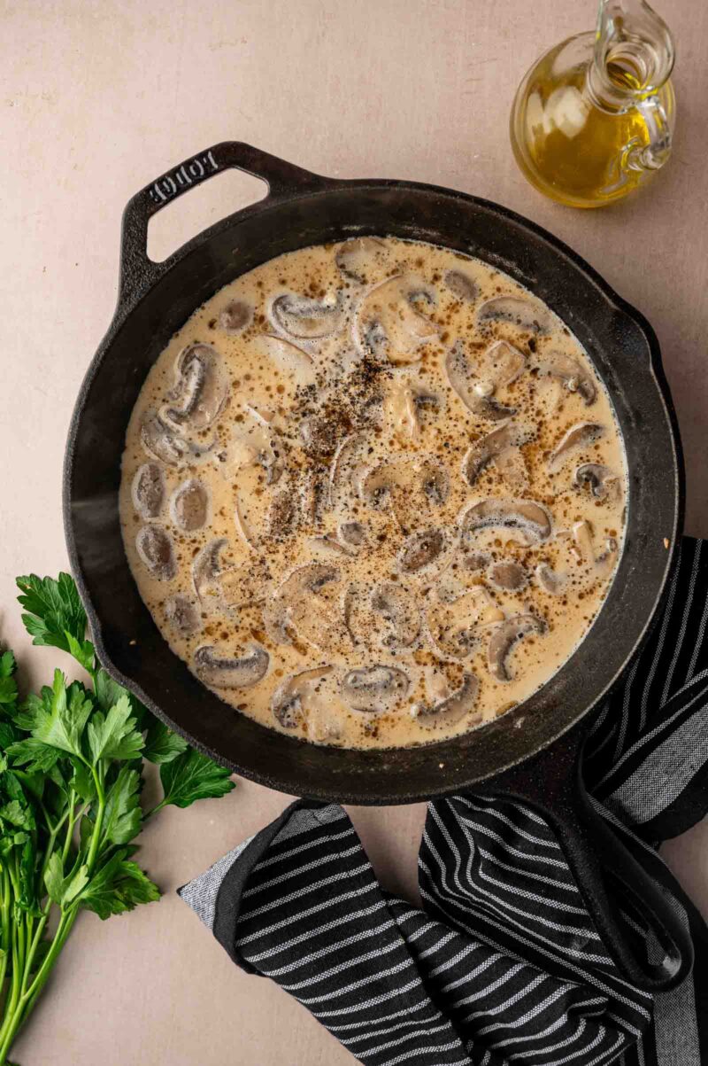 A cast iron skillet filled with creamy marsala sauce, topped with ground pepper, sits on a counter beside fresh parsley, a black striped towel, and a bottle of olive oil.