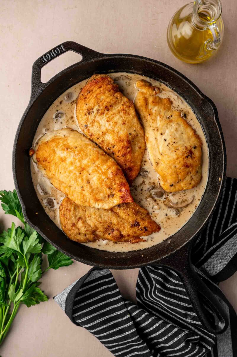 Four cooked chicken breasts in creamy marsala sauce in a black skillet, with a bunch of parsley, striped towel, and a glass bottle of oil nearby.