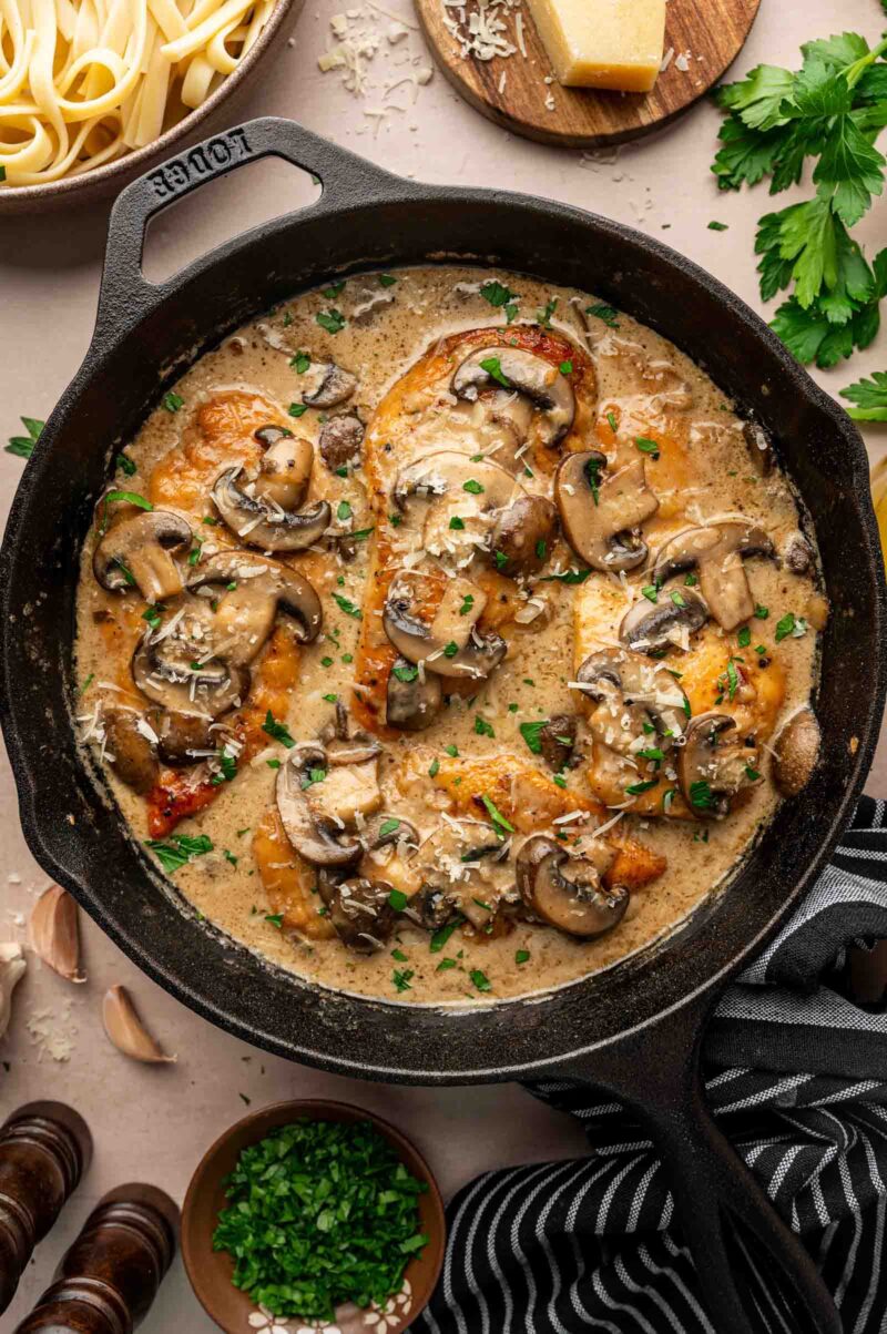 Chicken breasts in a creamy mushroom sauce are cooked in a black cast iron skillet, garnished with chopped parsley. Parsley, parmesan, garlic, and pasta are visible nearby.