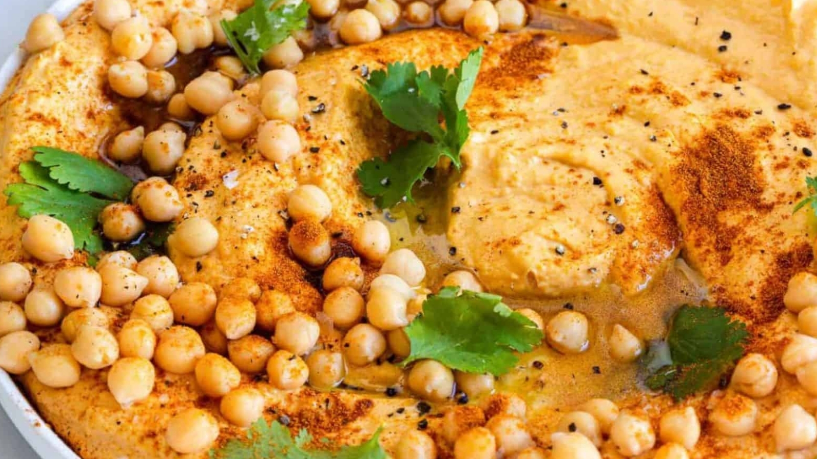 A close-up of a bowl of hummus topped with chickpeas, olive oil, paprika, black pepper, and cilantro leaves. The hummus has a creamy texture and is visually garnished with spices and herbs.