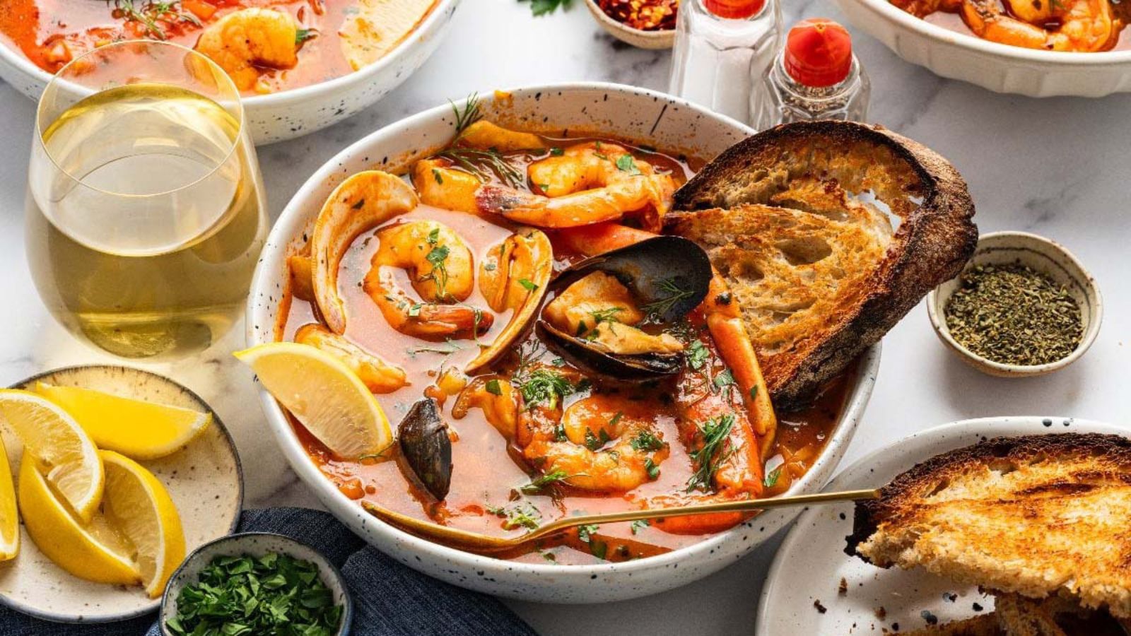 A bowl of seafood stew with shrimp, mussels, and toast sits on a table next to a glass of white wine, lemon wedges, and seasonings.