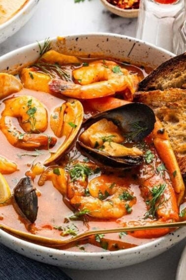 A bowl of seafood stew with shrimp, mussels, and toasted bread, garnished with herbs and lemon wedges, next to a glass of white wine.