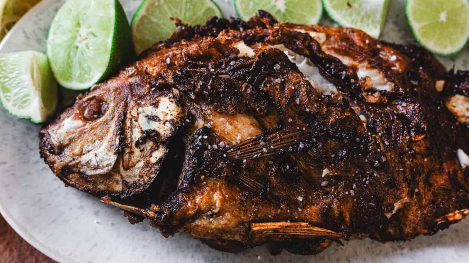 A whole fried fish with crispy, browned skin is served on a plate with several lime wedges arranged around it.