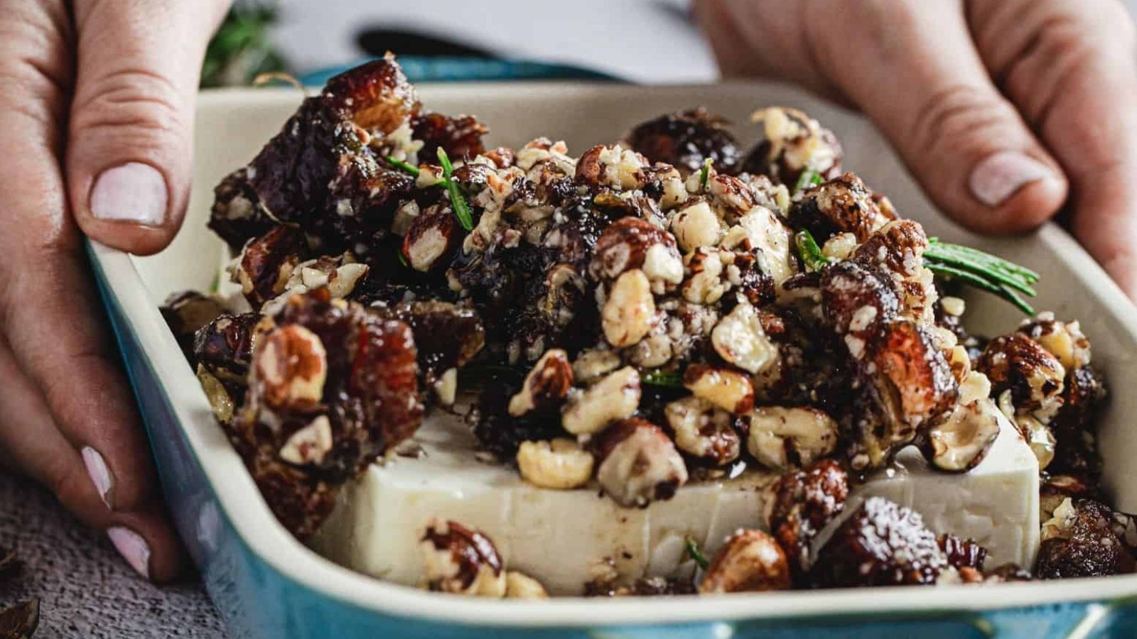 A block of white cheese in a rectangular baking dish is topped with chopped nuts and dates, with hands holding the dish on either side.