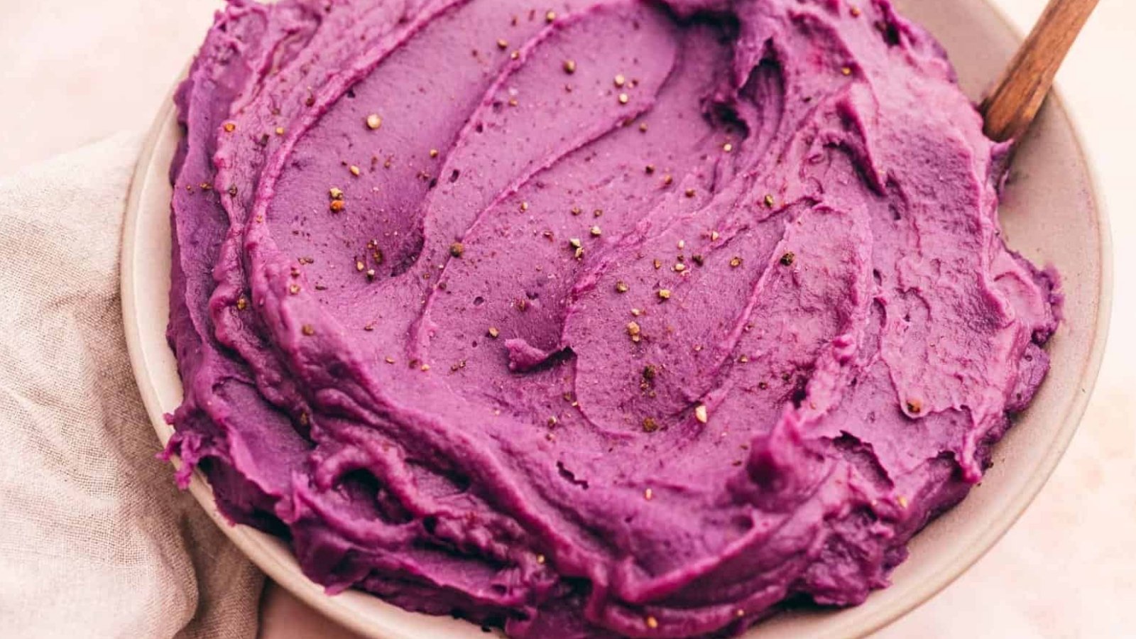 A bowl of smooth purple mashed potatoes with a wooden spoon, sprinkled with black pepper.