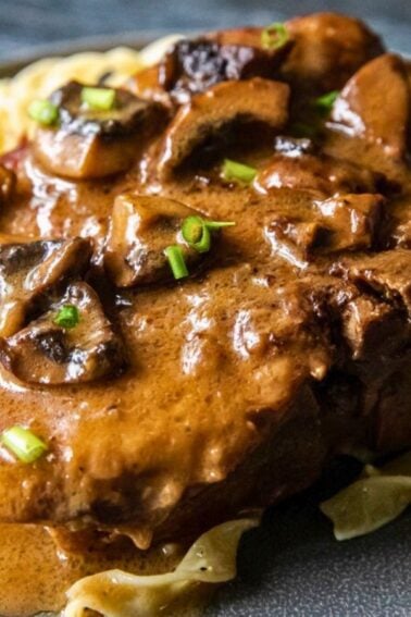 A plate of egg noodles topped with a slice of beef covered in brown mushroom gravy and garnished with chopped green herbs.