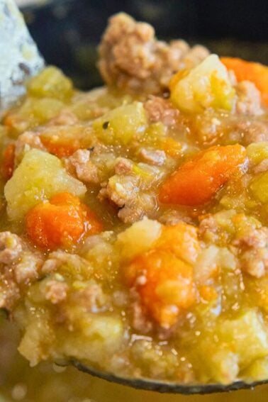 A ladle holding a thick soup or stew with chunks of carrots, potatoes, ground meat, and a greenish broth, held over a pot.