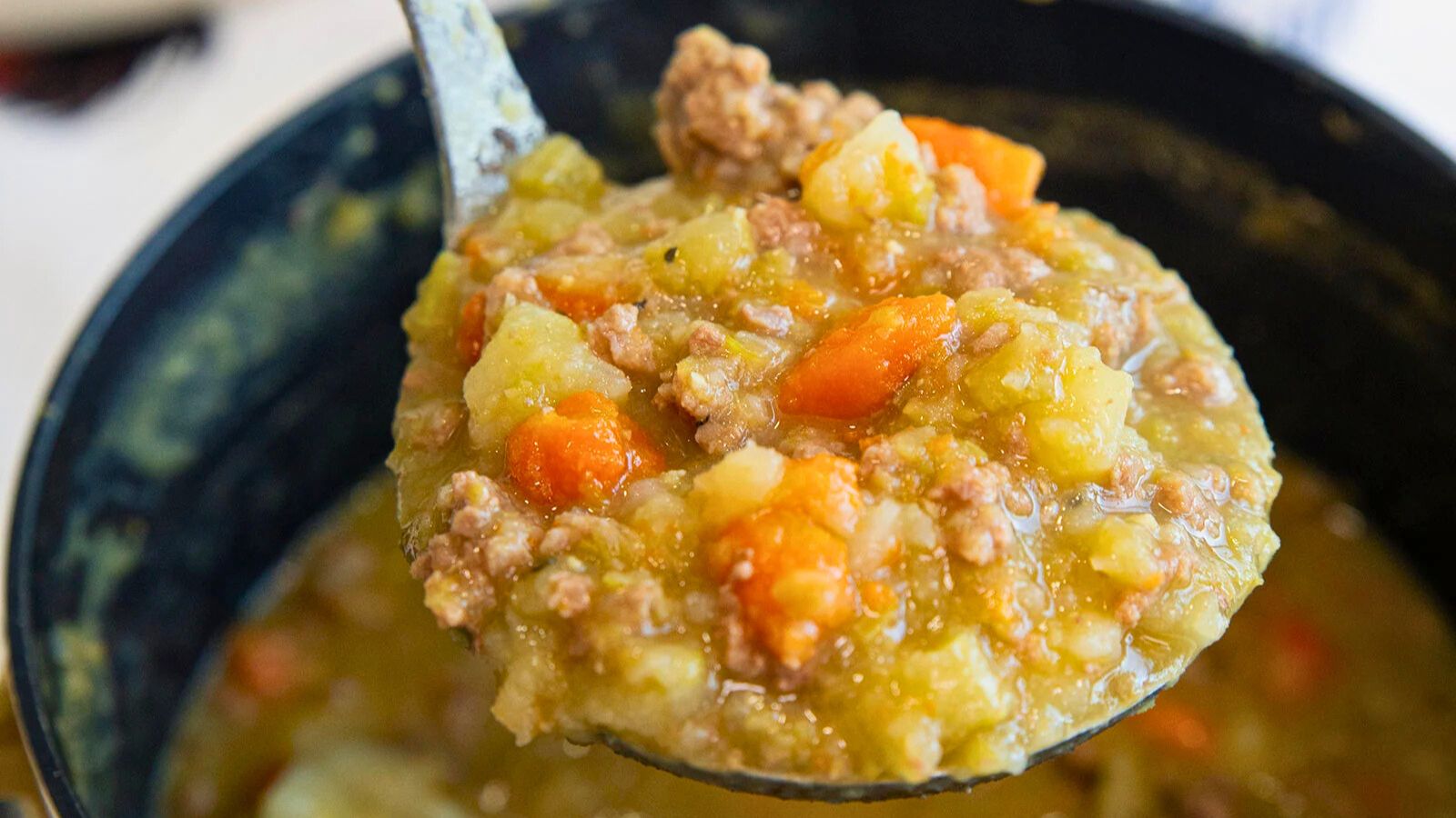 A ladle holding a thick soup or stew with chunks of carrots, potatoes, ground meat, and a greenish broth, held over a pot.