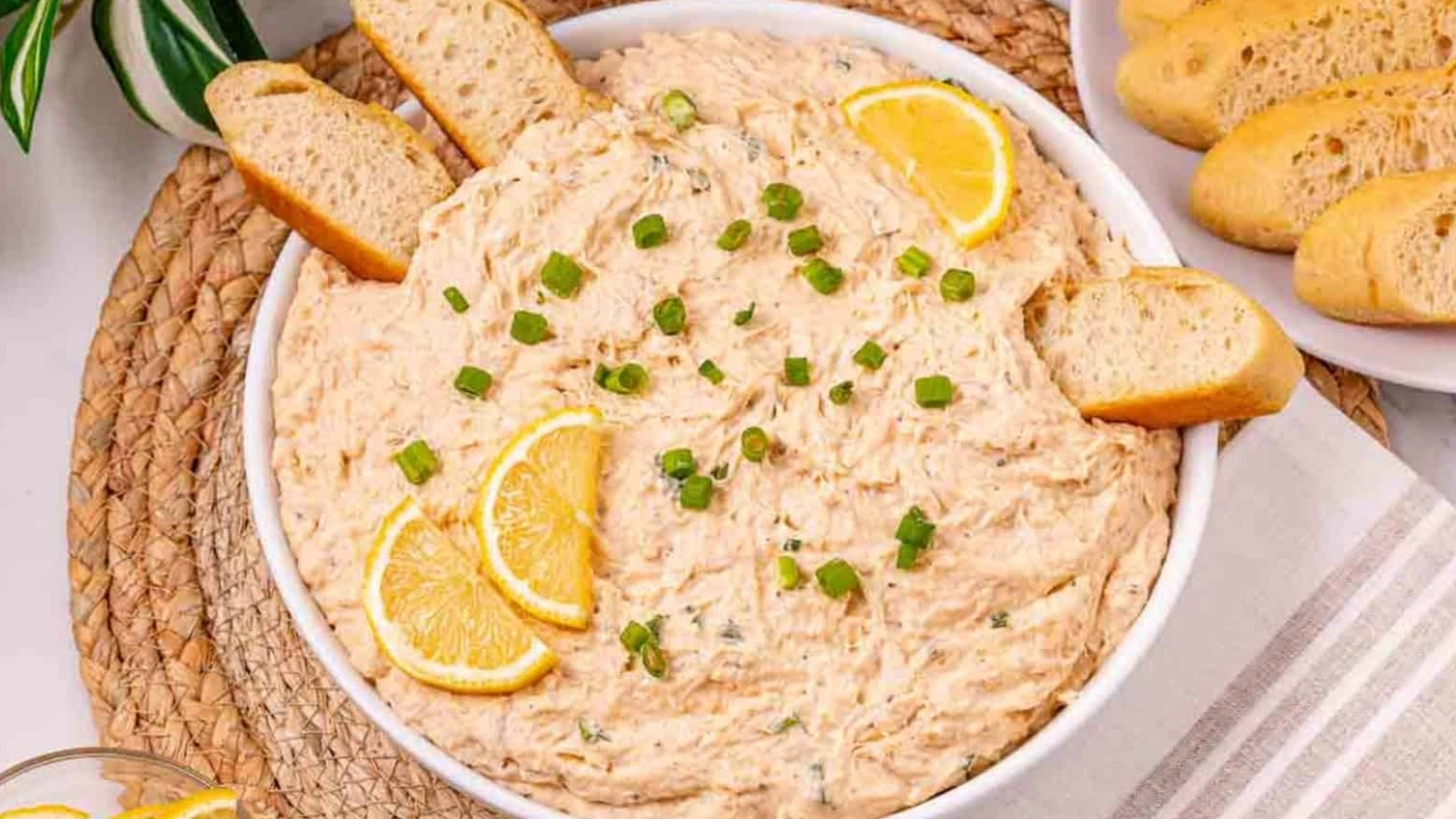 A bowl of creamy dip garnished with lemon slices and green onions, served with slices of baguette.