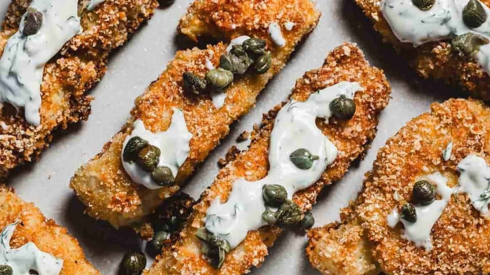 Golden breaded fish sticks topped with creamy sauce and capers, arranged on a light-colored surface.
