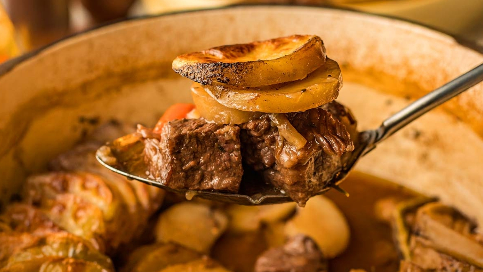 A spoon holds chunks of beef topped with potato slices above a pot containing broth, potatoes, carrots, and pieces of meat.