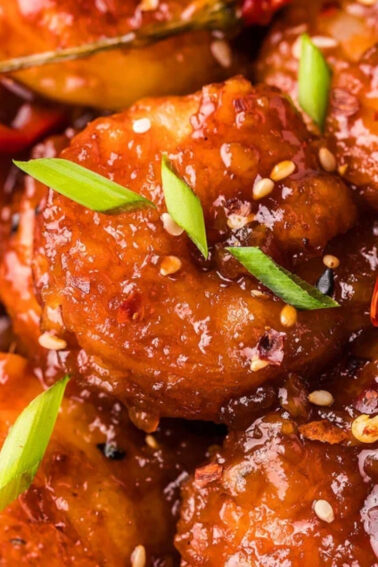 Close-up of glazed, saucy shrimp garnished with sliced green onions, red chili peppers, and sesame seeds, served over white rice.