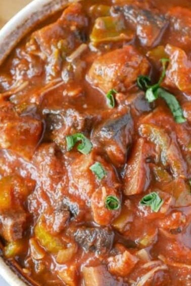 A bowl of chunky tomato-based stew with pieces of meat and vegetables, garnished with fresh herbs. Bread, cheese, and basil leaves are nearby on a wooden board.