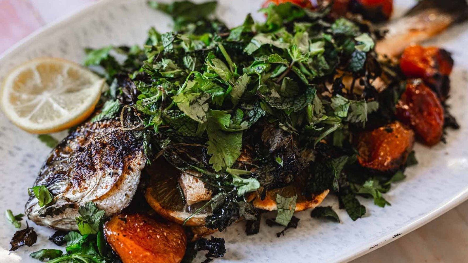 A whole roasted fish topped with chopped herbs and lemon slices is served on a white platter with roasted tomatoes and greens.