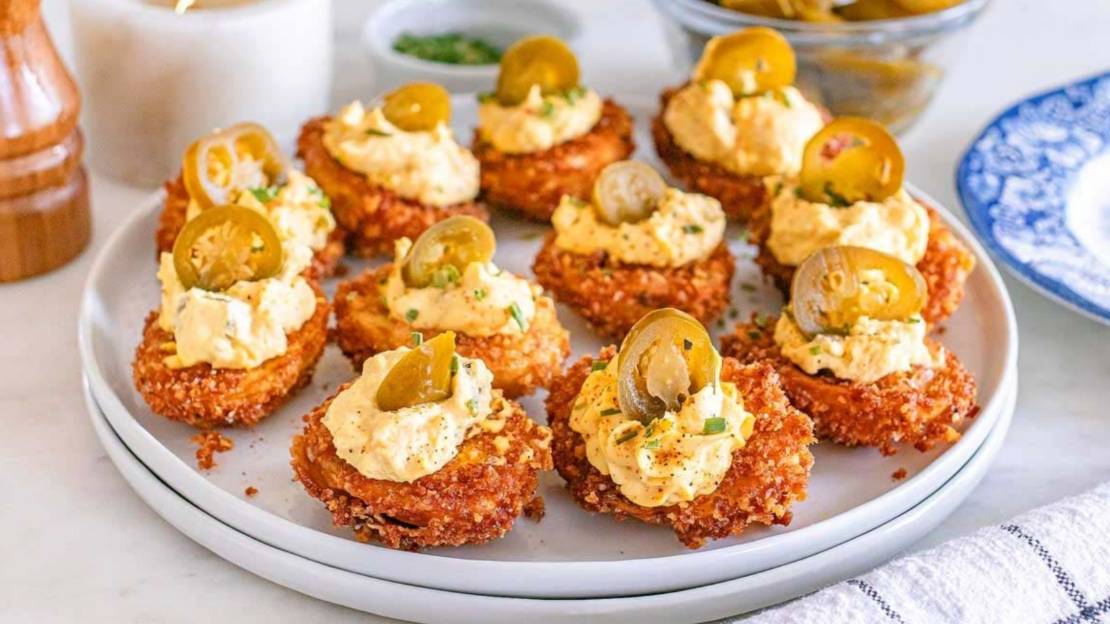 Round fried potato slices topped with a dollop of creamy filling and sliced jalapeños are arranged on a plate with bowls and a candle in the background.