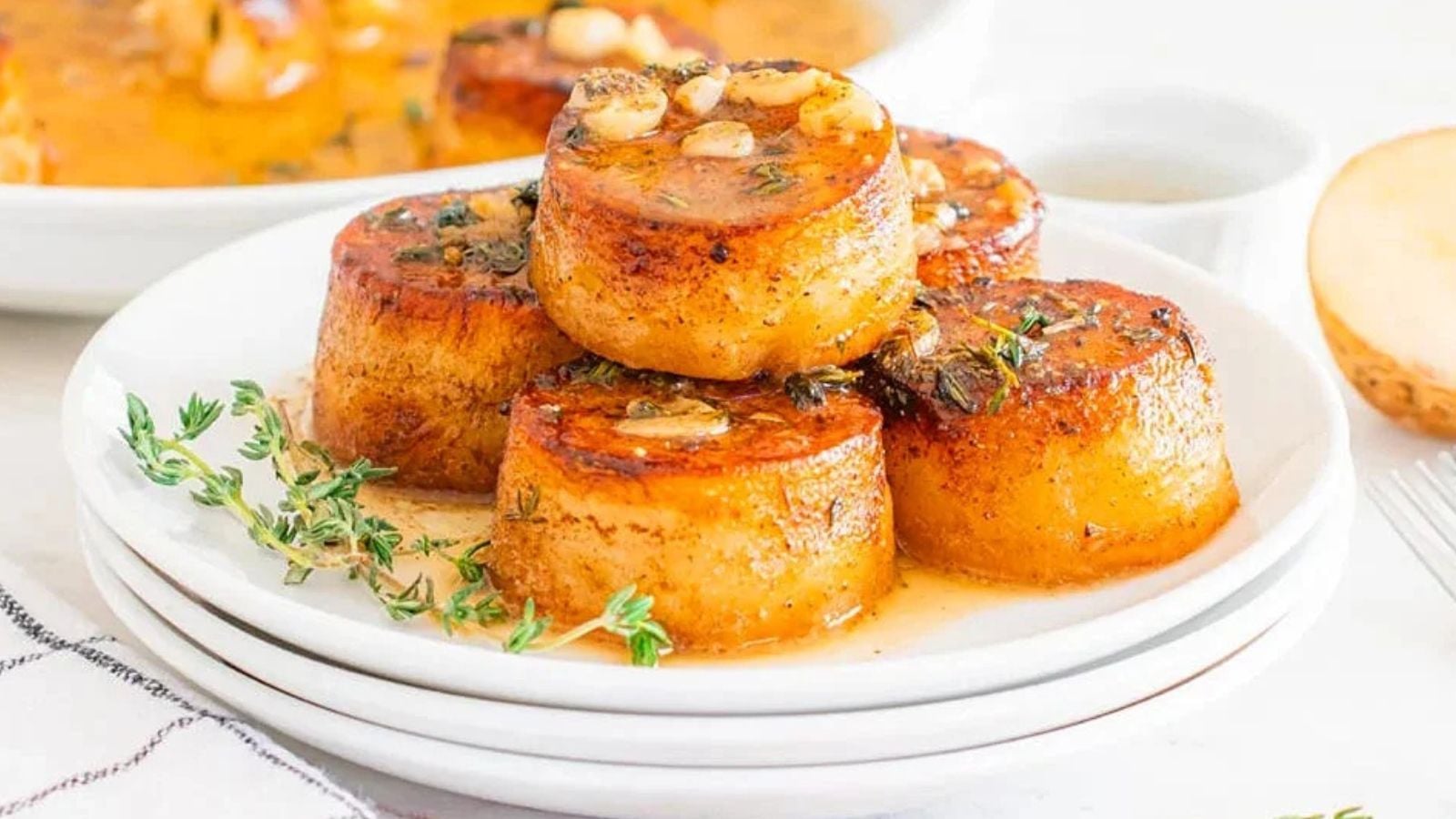 Five browned fondant potatoes garnished with herbs and garlic sit stacked on a white plate, with sauce pooling at the bottom and a sprig of thyme on the side.