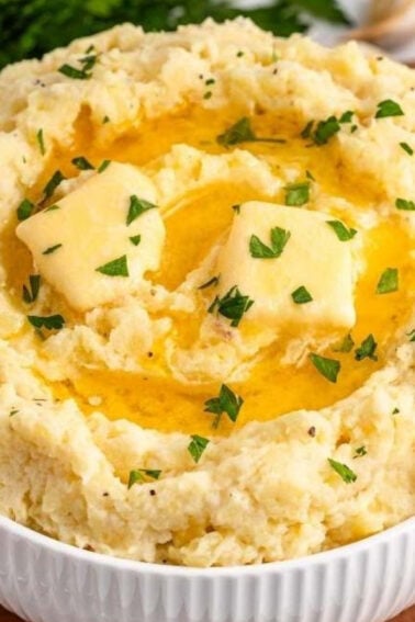 A bowl of mashed potatoes topped with melting butter and chopped herbs, placed on a wooden surface with garlic and parsley in the background.