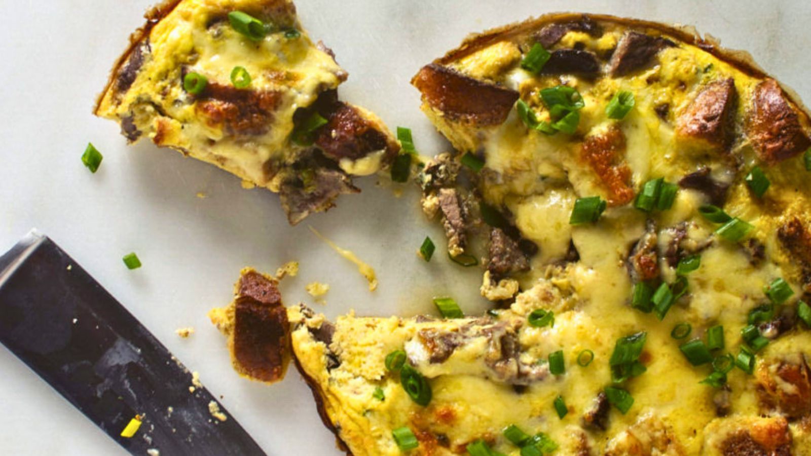 A sliced frittata with mushrooms and chopped green onions on a light surface, with a serving spatula next to it.