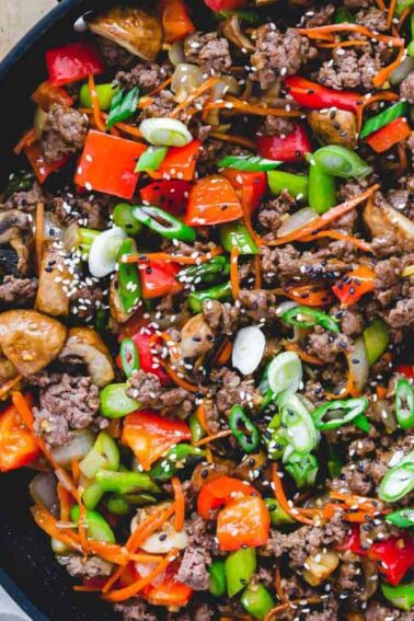 A skillet filled with a colorful stir-fry of ground meat, red bell peppers, mushrooms, carrots, green onions, and sesame seeds.