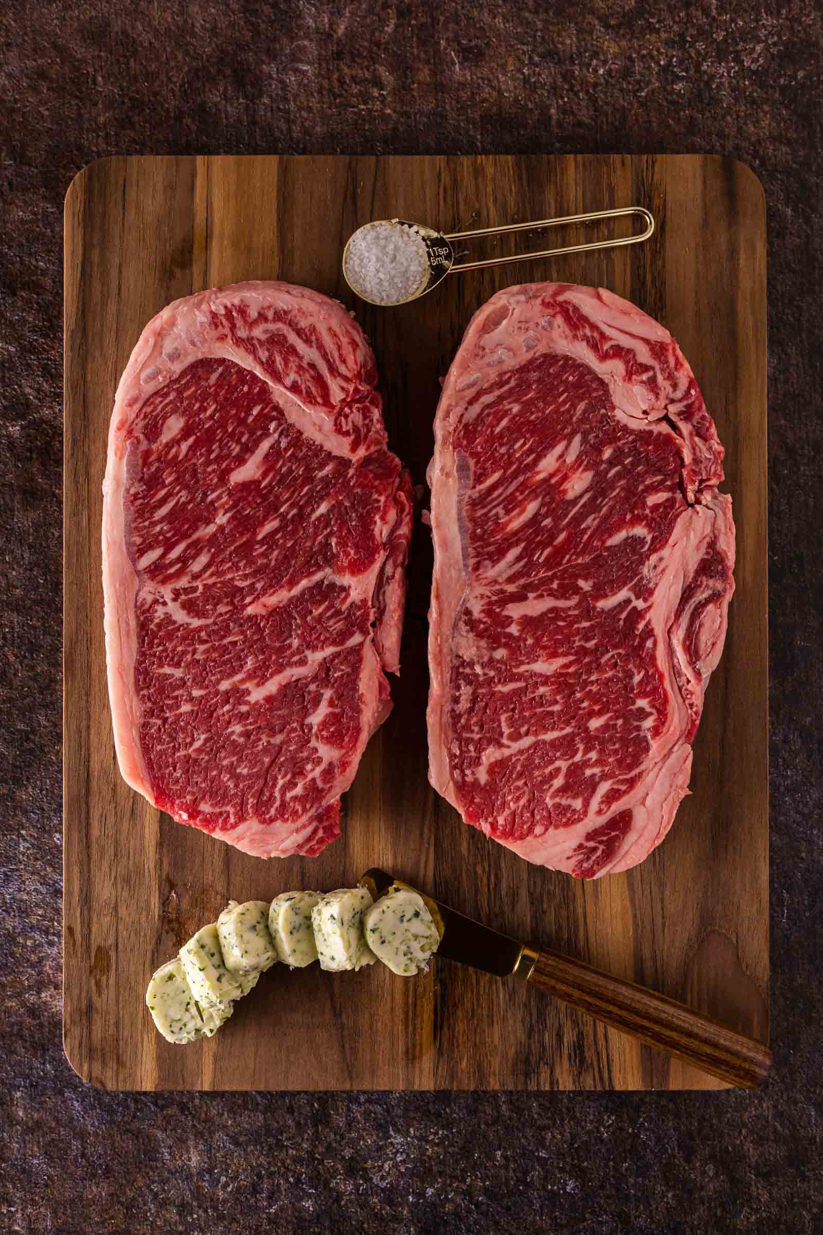 Two raw ribeye steaks on a wooden cutting board with a spoonful of coarse salt and several slices of herb butter, plus a knife.