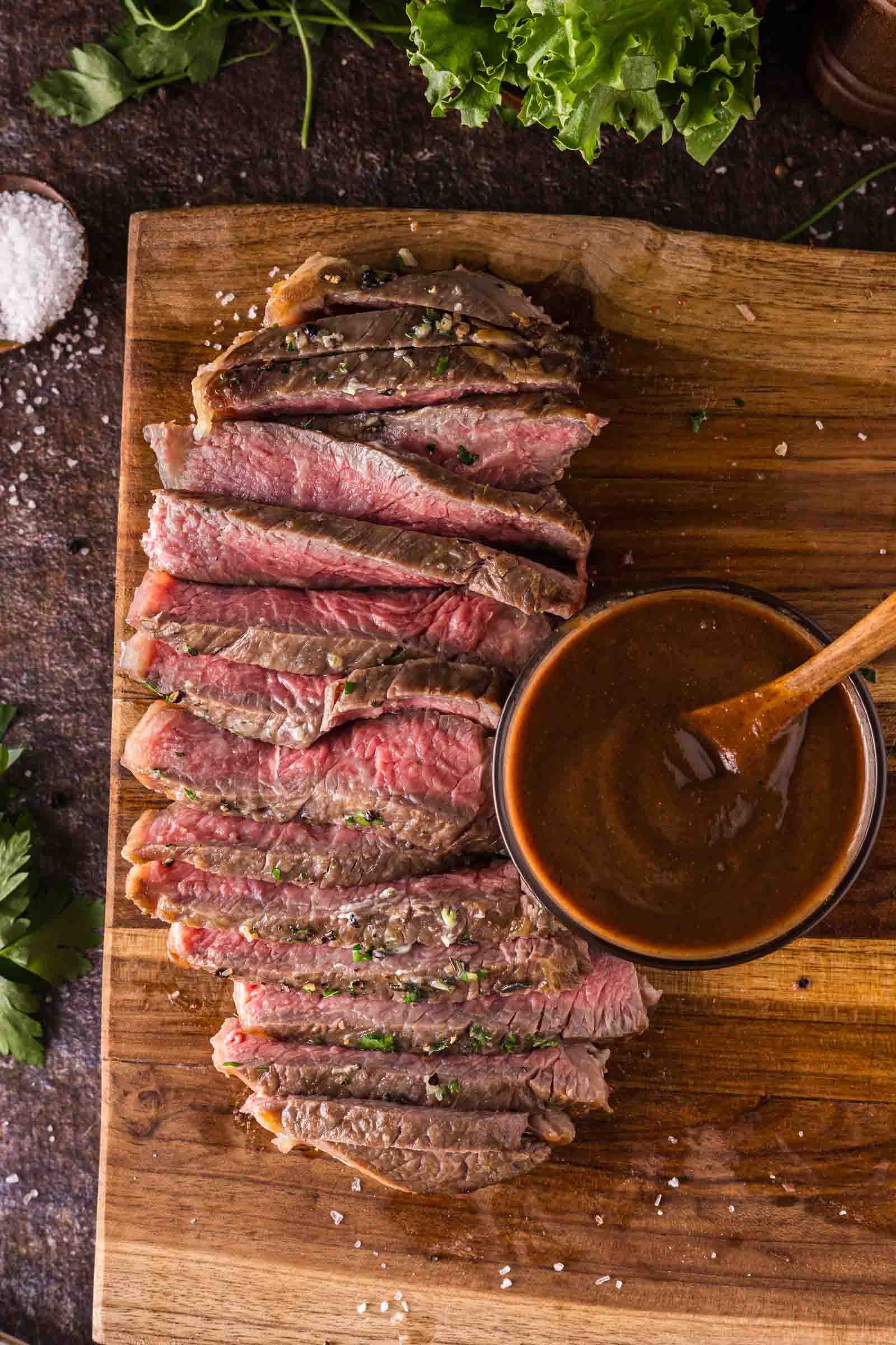 Sliced medium-rare steak arranged on a wooden cutting board with a round dish of steak sauce and a wooden spoon.