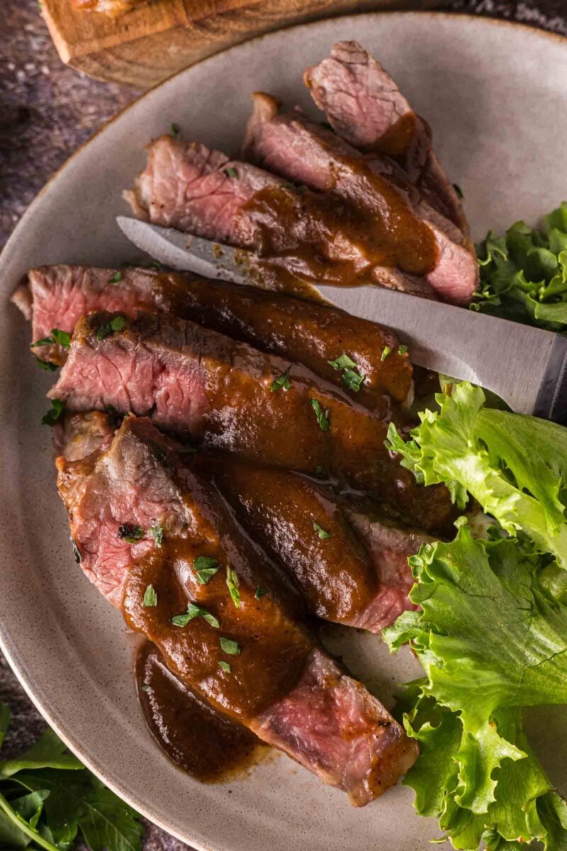 Sliced steak with a1 sauce on a beige plate, garnished with chopped herbs and served with leafy greens.