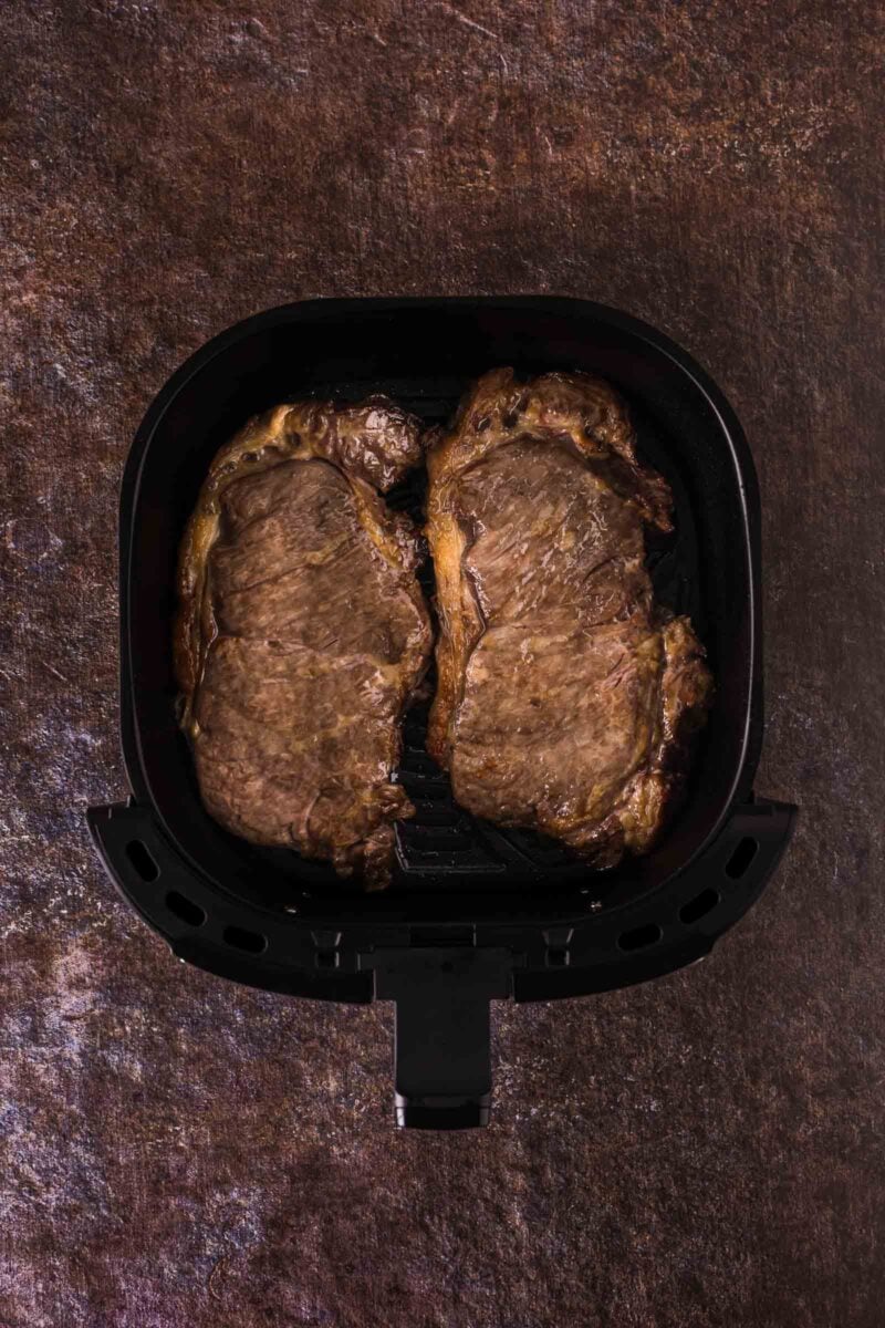 Two cooked steaks are placed side by side inside the basket of an air fryer, viewed from above against a dark textured background.