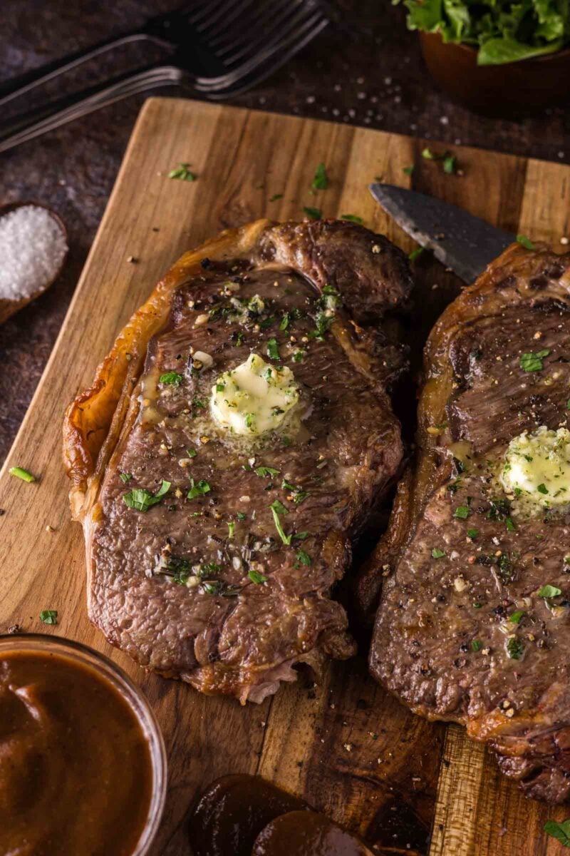 Two air fried steaks topped with melting butter and chopped herbs sit on a wooden cutting board, with a knife, sauce, and utensils nearby.