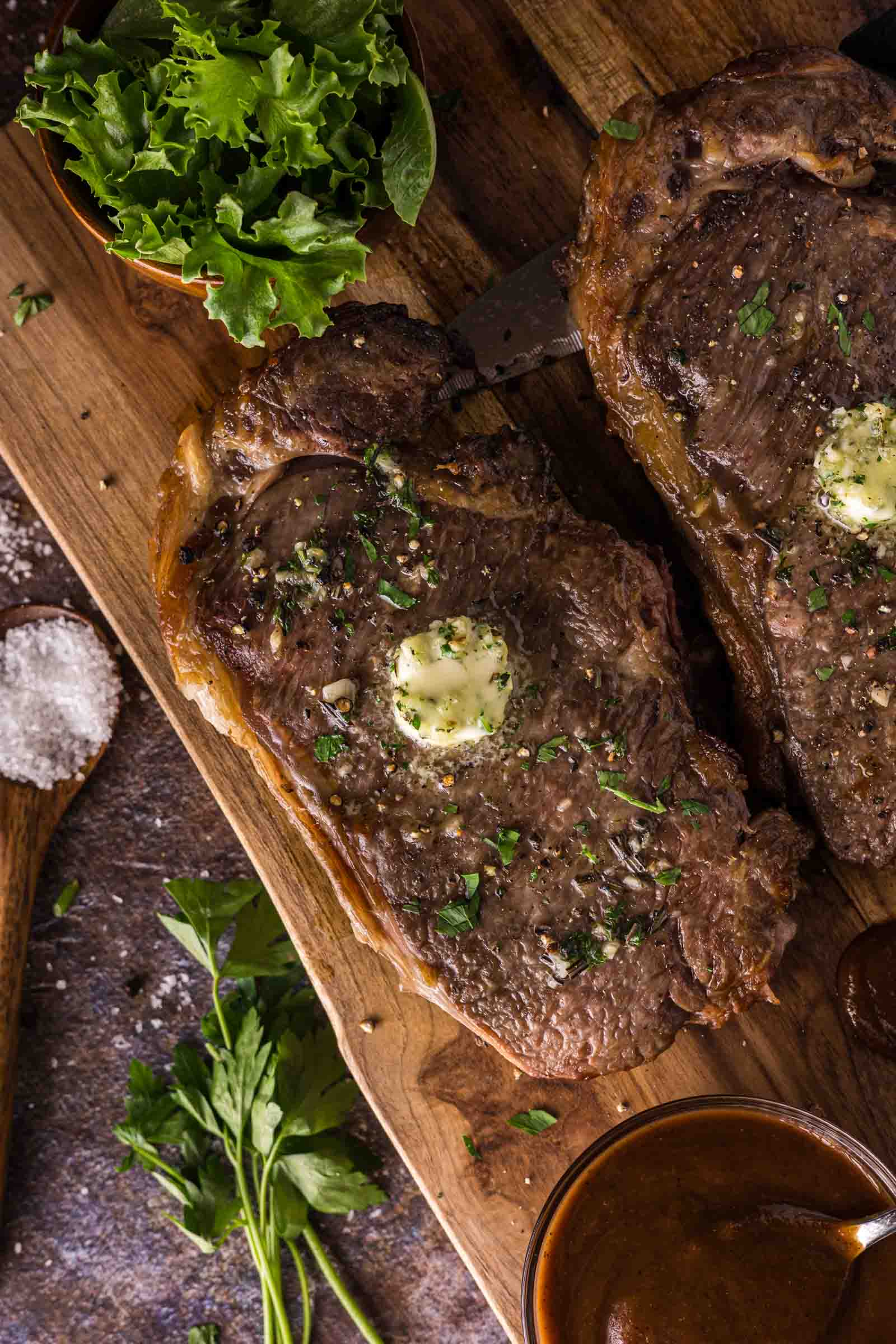 An air fried ribeye steak topped with a pat of butter sits on a wooden board, surrounded by fresh greens, salt, and a small bowl of sauce.