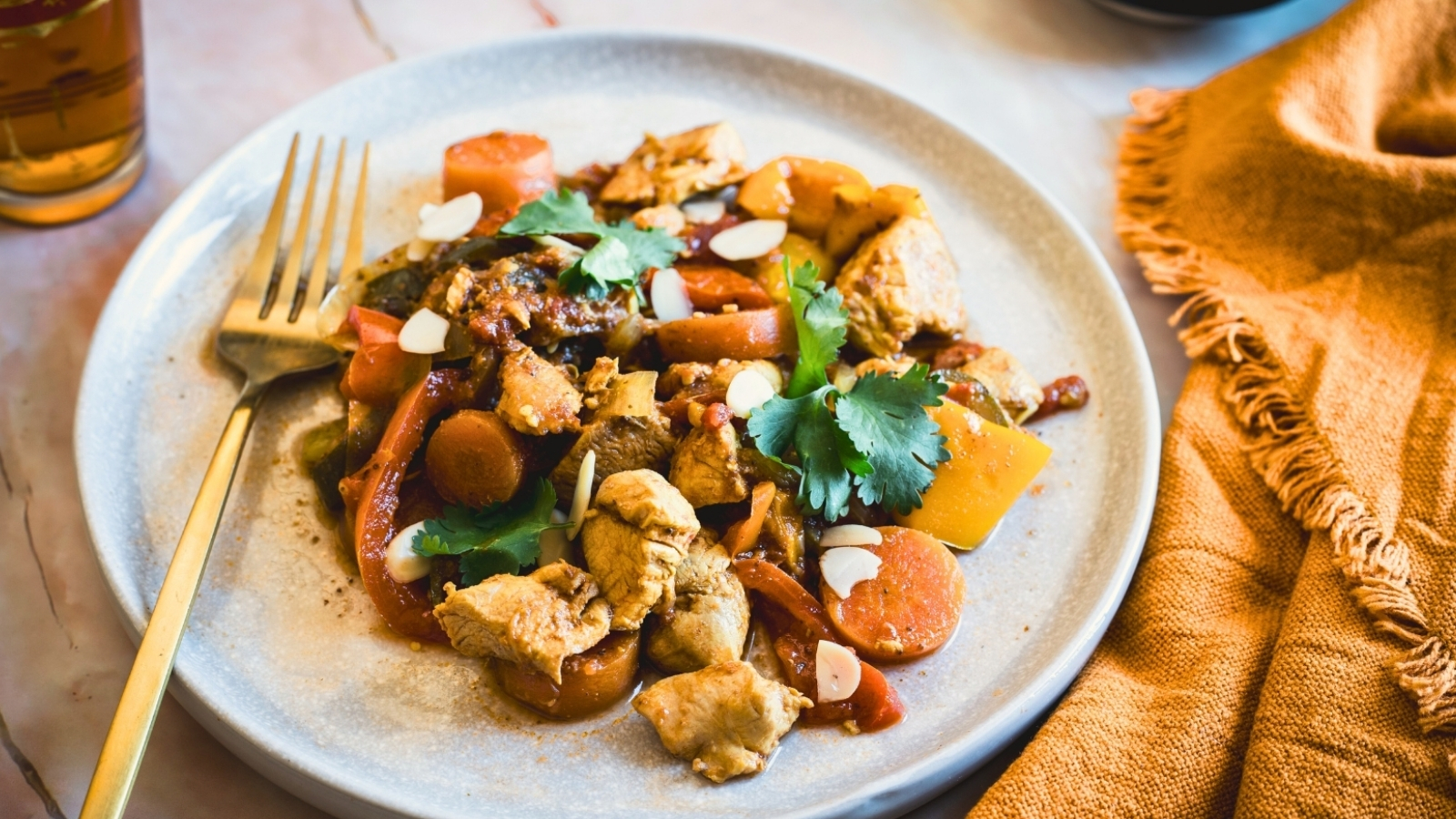 A plate of chicken stir-fry with sliced carrots, bell peppers, and mushrooms, garnished with cilantro and almond slices. A fork rests on the plate, and a yellow cloth is visible in the background.