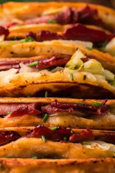 A row of crispy tacos filled with sliced corned beef and shredded cabbage, garnished with chopped chives, arranged on a rectangular white plate.