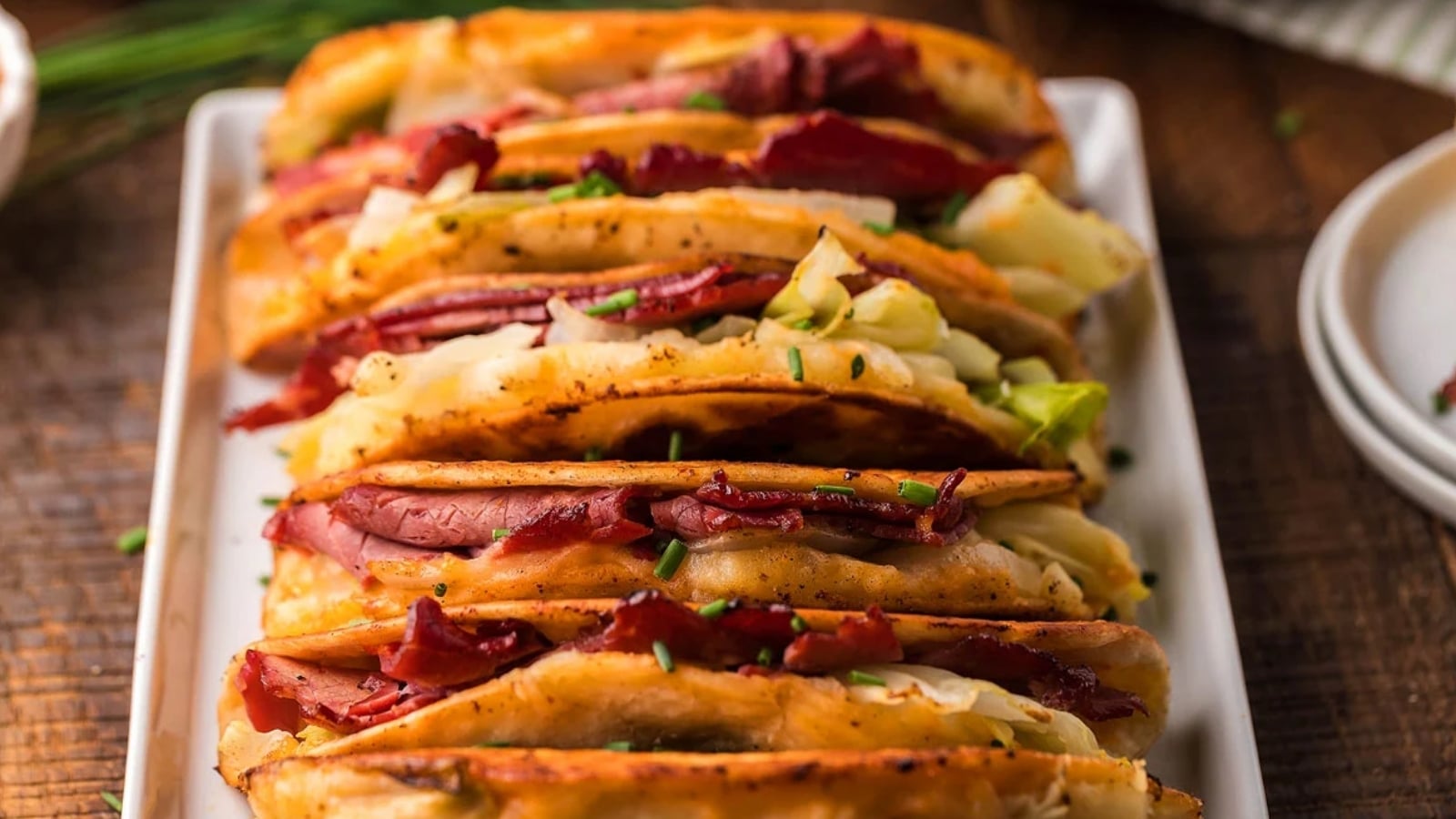 Several folded tacos filled with sliced meat, bacon strips, melted cheese, and chopped green onions are arranged in a row on a rectangular white plate.