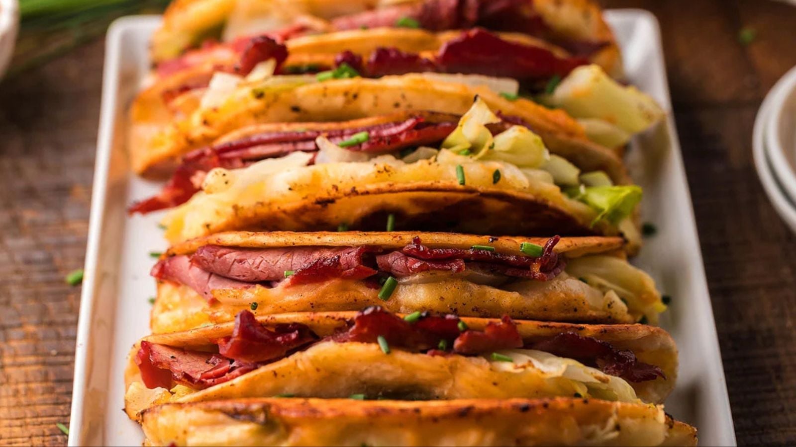 A row of crispy tacos filled with sliced corned beef, cabbage, and chopped chives on a rectangular white platter.