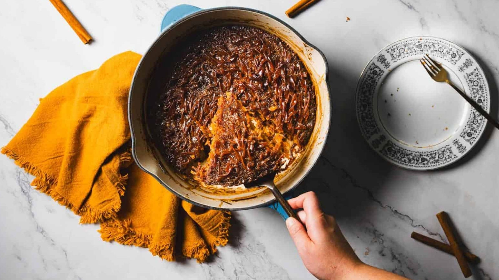 A hand scoops baked kugel from a round dish on marble, beside an empty plate, fork, orange napkin, and cinnamon sticks.