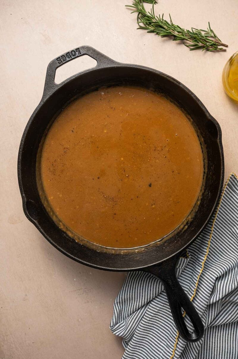 A cast iron skillet filled with brown gravy sits on a light surface next to a sprig of rosemary, a striped cloth, and a partial glass container of oil.