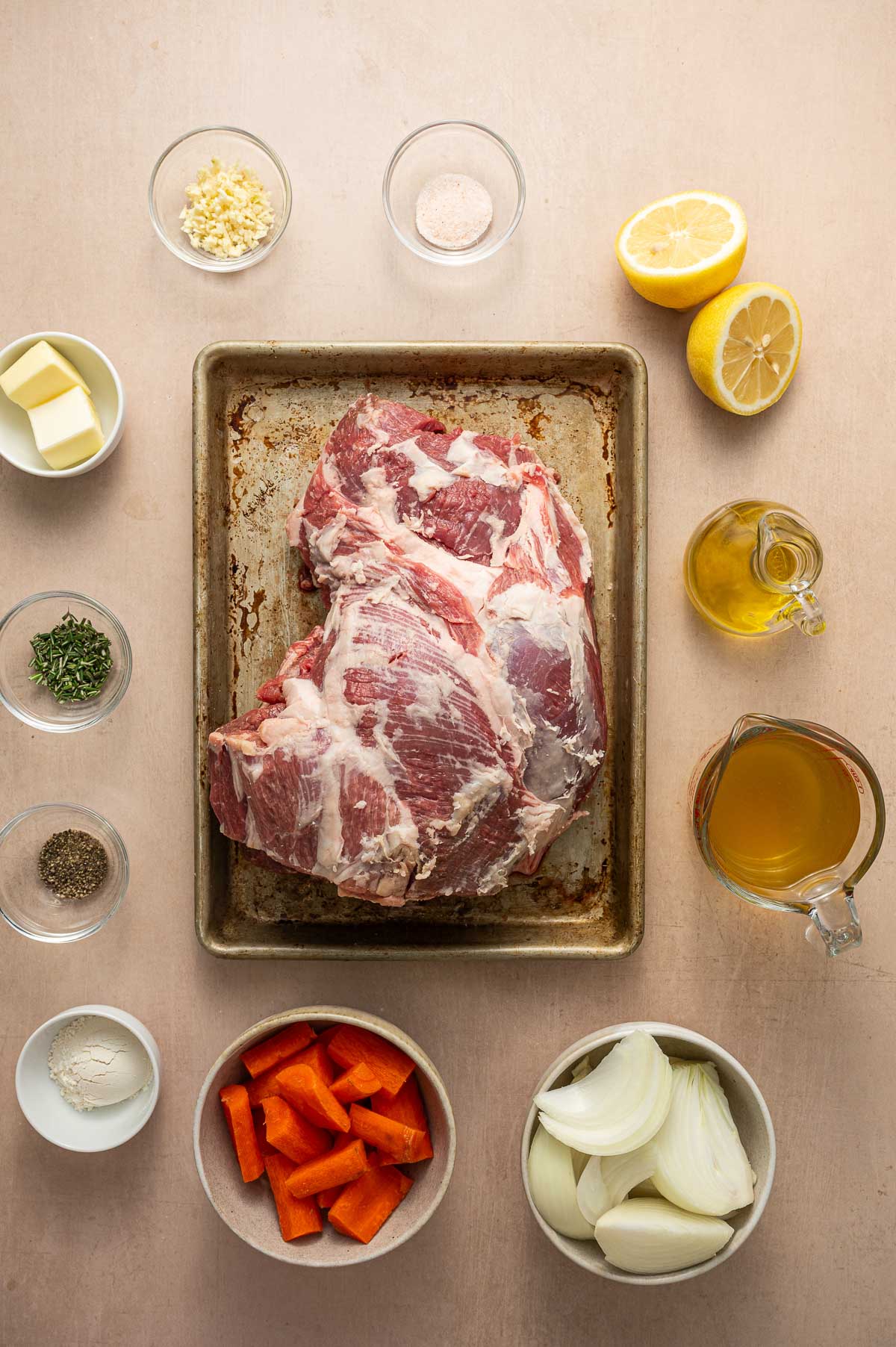 Raw boneless leg of lamb on a baking sheet surrounded by bowls of chopped carrots, onions, garlic, butter, spices, salt, broth, olive oil, and a halved lemon on a beige surface.