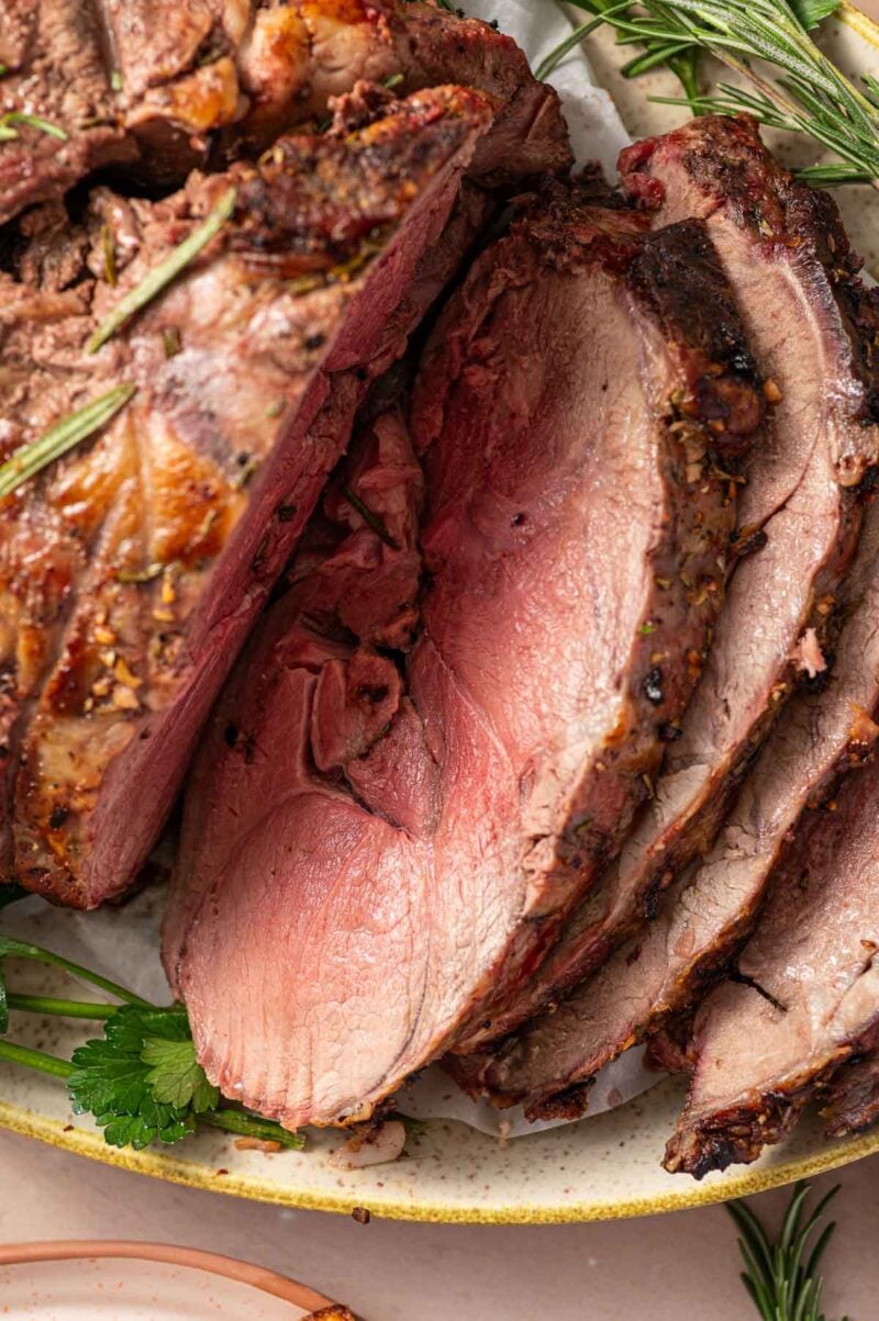 Sliced roast lamb shown on a plate, cooked medium rare, garnished with fresh rosemary and parsley.