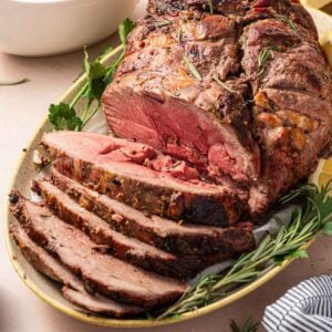 A sliced roast lamb garnished with rosemary and parsley on a platter, with a gravy boat and lemon wedges in the background.