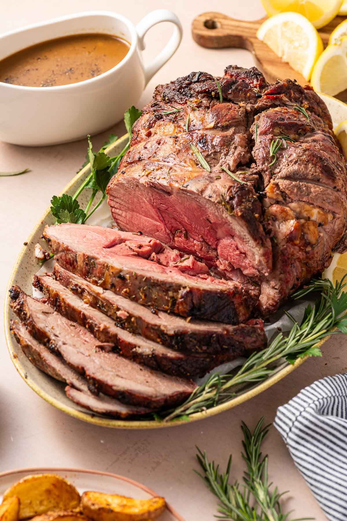 A sliced roast lamb garnished with rosemary and parsley on a platter, with a gravy boat and lemon wedges in the background.