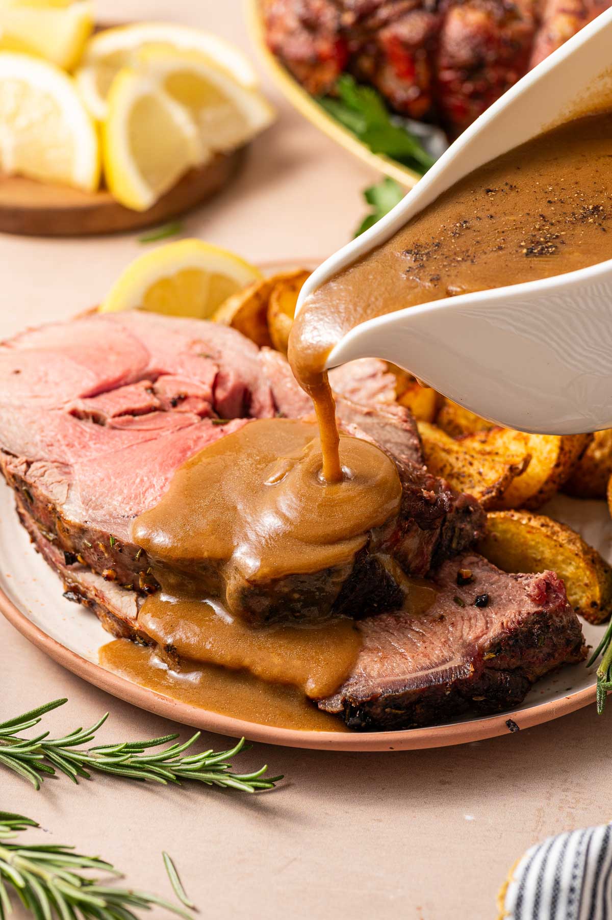 Gravy being poured over slices of roast lamb on a plate with roasted potatoes and lemon wedges, with rosemary garnish nearby.