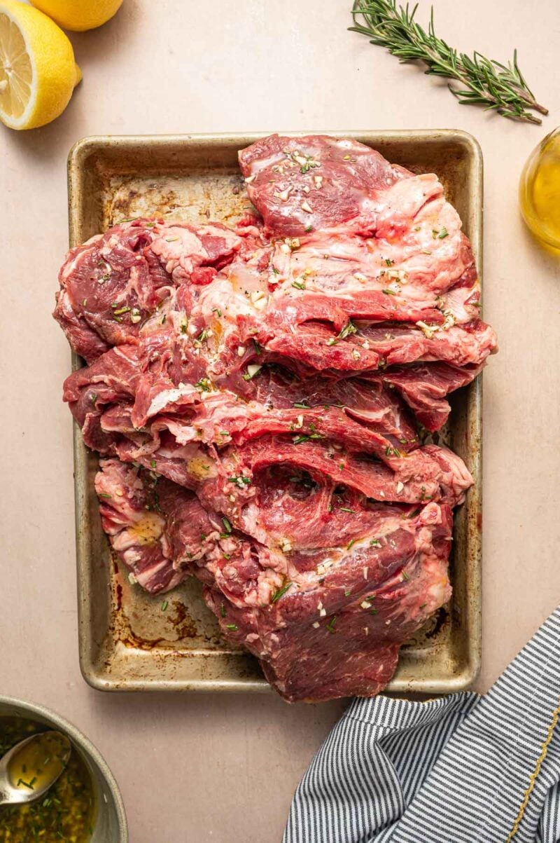 Raw season boneless leg of lamb butterflied open on a baking tray, surrounded by lemon halves, rosemary, olive oil, and a striped kitchen towel.