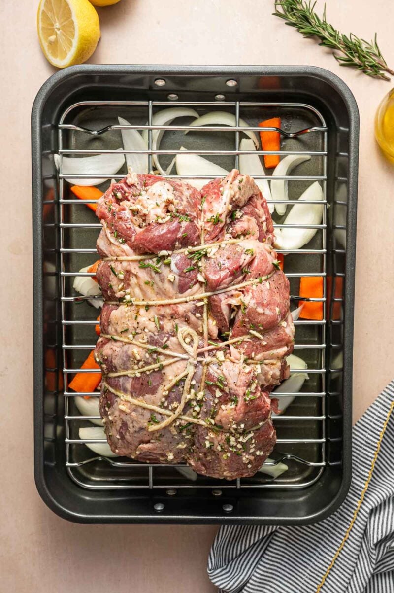 Uncooked, seasoned, and tied lamb roast sits on a rack over chopped onions and carrots in a roasting pan, with a lemon, rosemary, and striped towel nearby.