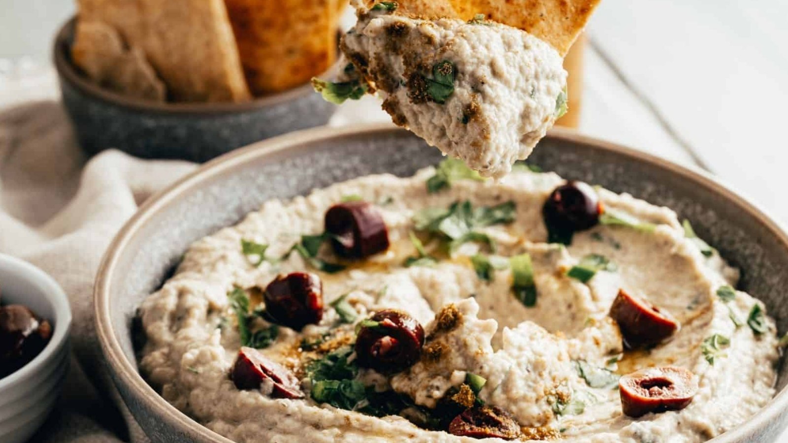 A bowl of baba ganoush topped with chopped herbs and olives, with a pita chip being dipped into the spread.