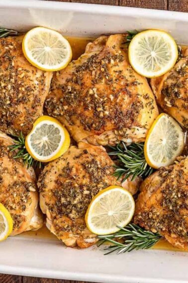 Baked chicken thighs in a white dish, garnished with lemon slices, rosemary sprigs, and herbs. Rice and green beans on plates nearby.