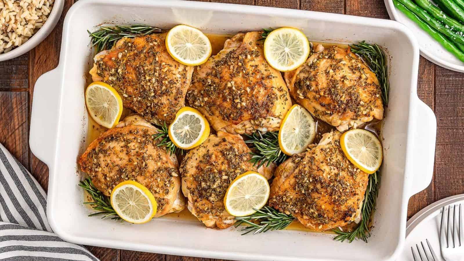 Baked chicken thighs in a white dish, seasoned with herbs and lemon slices, garnished with rosemary sprigs.